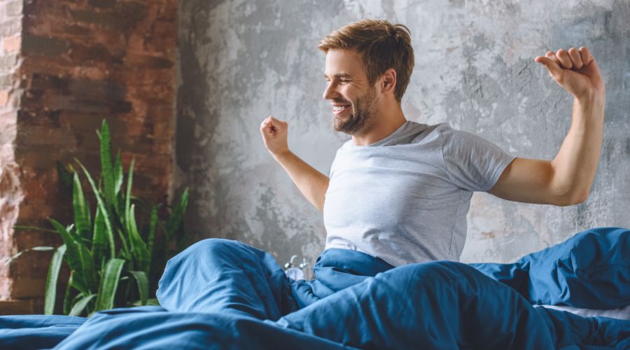 smiling young man waking up in bed and stretching his arms